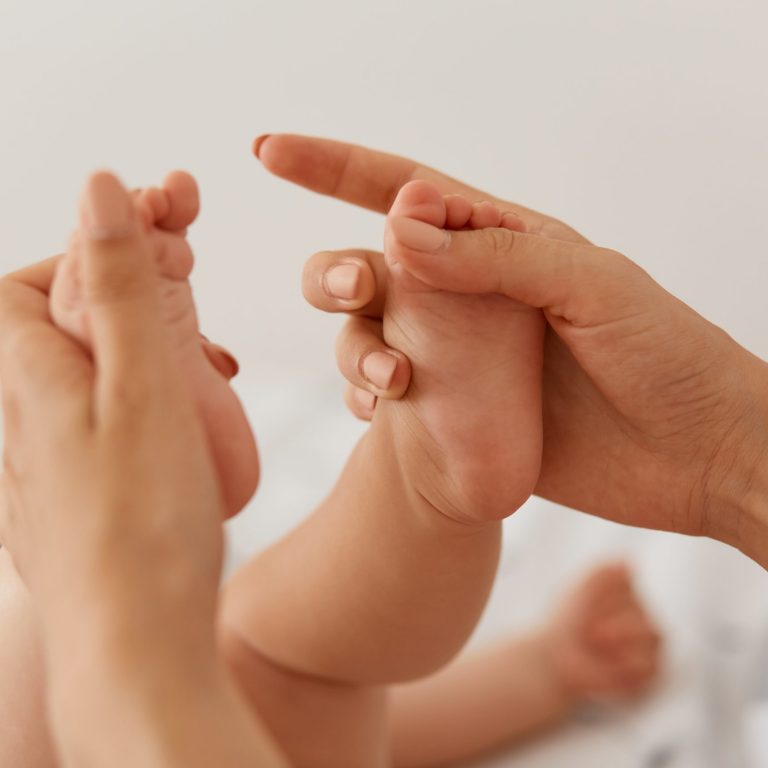 Close up portrait of baby legs in mother's hands, mommy makinng massage for her newborn baby against light room wall, woman arms touching infants body.