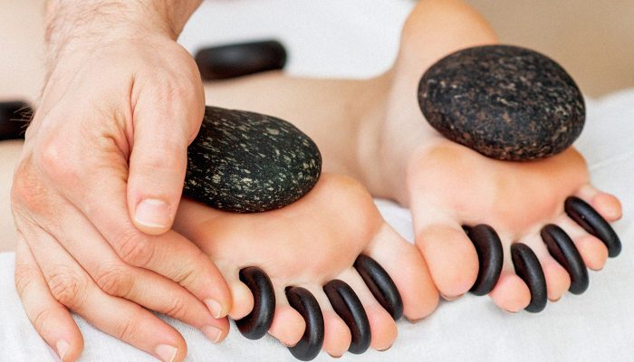 Hands of masseur putting hot little black stones between female toes for feet massage